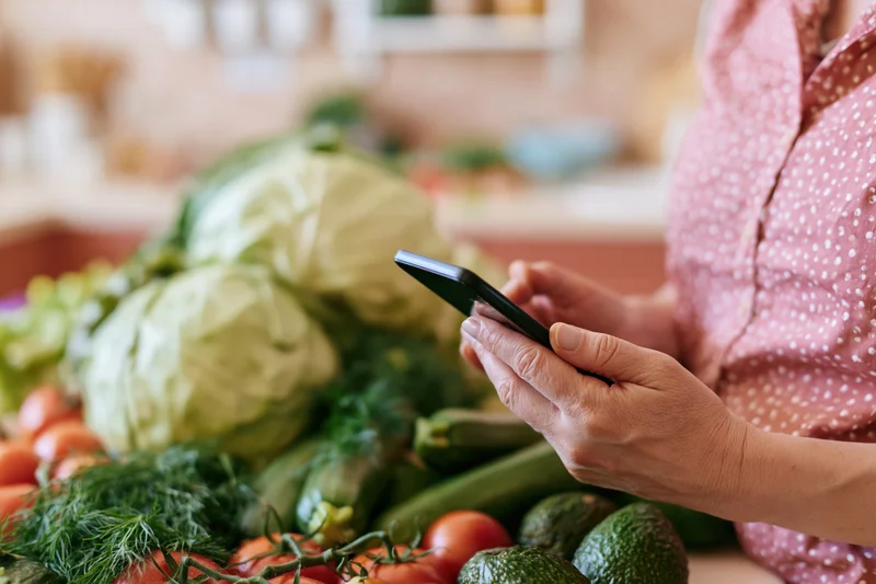 Woman using smartphone to browse recipe or grocery list beside fresh vegetables on kitchen counter