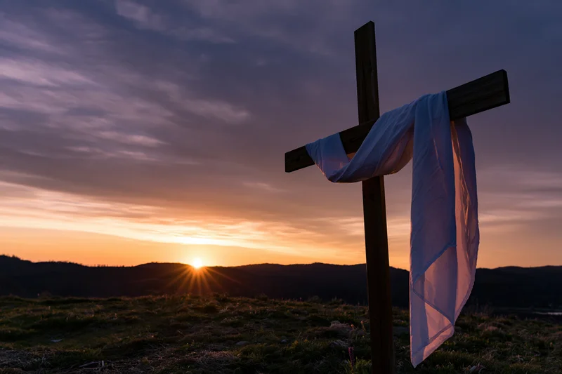 Wooden cross with white cloth draped over it standing on a hill at sunset