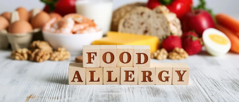 Wooden blocks spelling food allergy arranged on a white surface with various food items in the background
