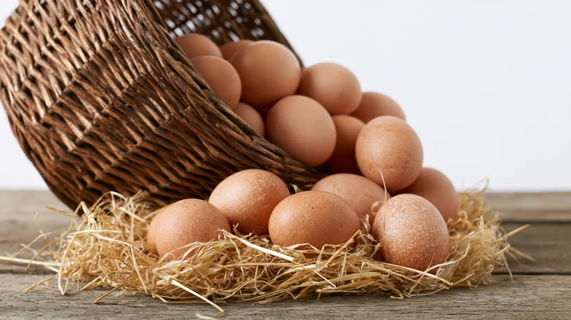 Brown chicken eggs spilling from a tipped wicker basket onto straw on a wooden table