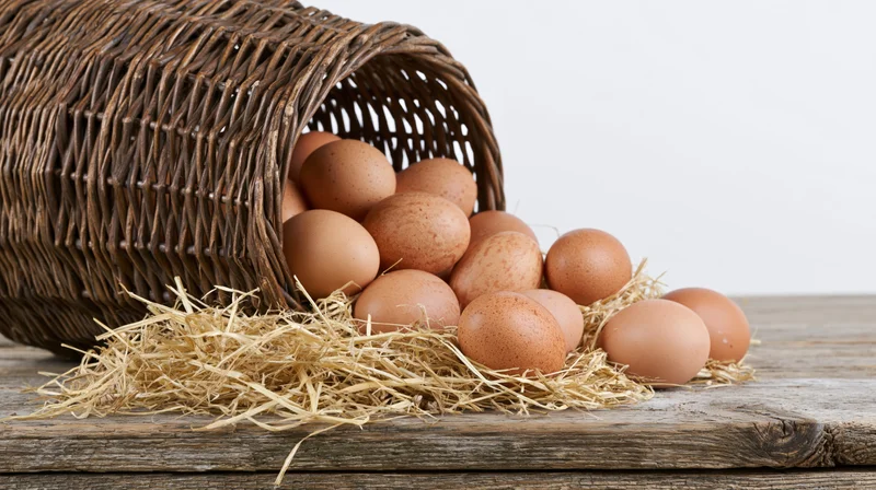 Brown chicken eggs spilling from a tipped wicker basket onto straw on a wooden table