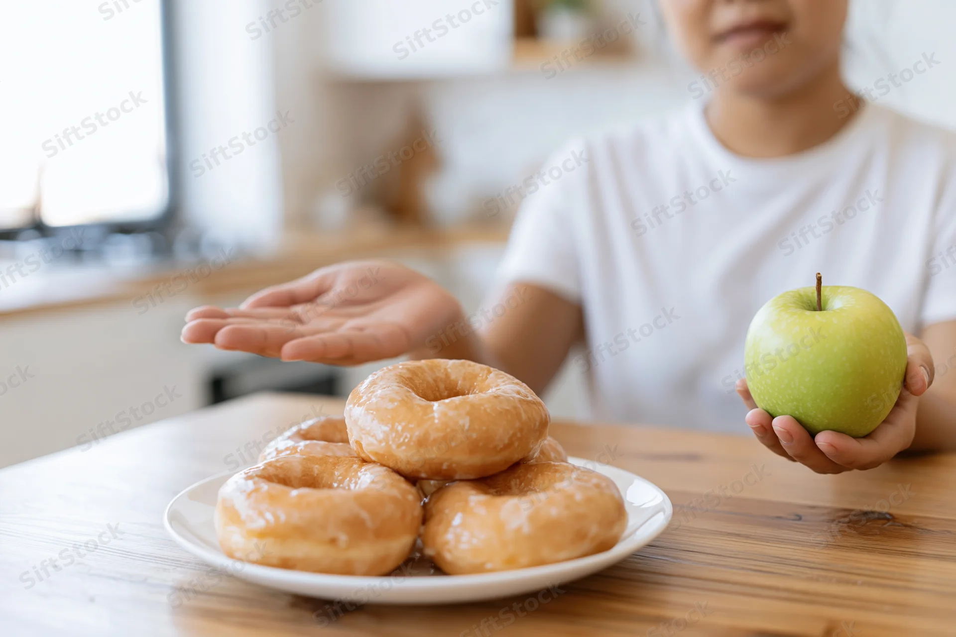 Woman holding green apple while pushing away plate of glazed donuts at kitchen table