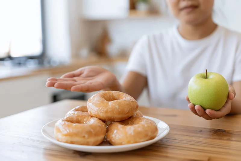 Woman holding green apple while pushing away plate of glazed donuts at kitchen table