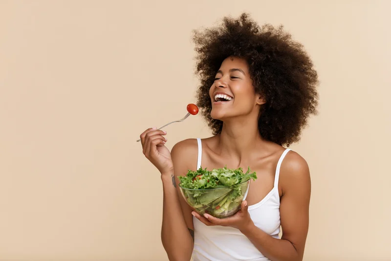 Young black woman laughing while eating a salad with a cherry tomato on a fork, holding a glass bowl against a beige background