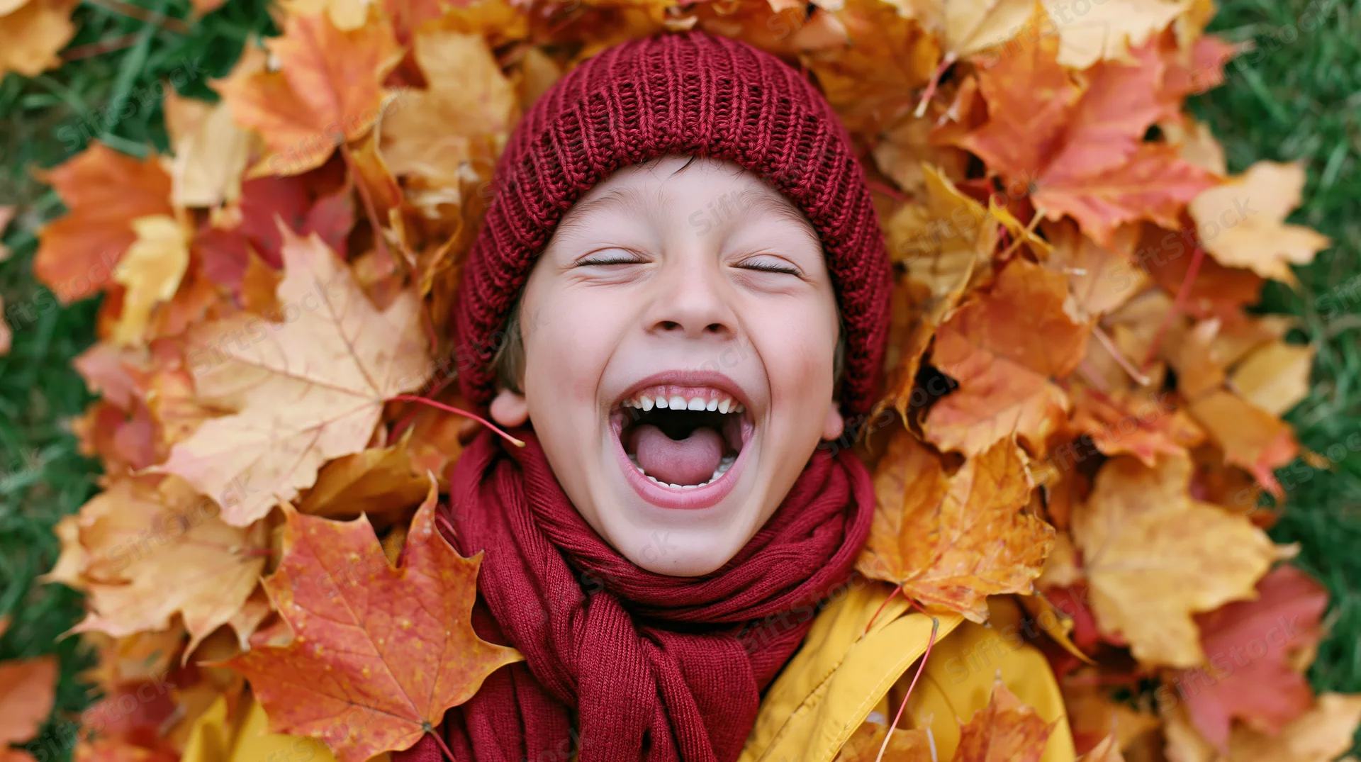 Boy laughing with eyes closed while buried in autumn maple leaves outdoors
