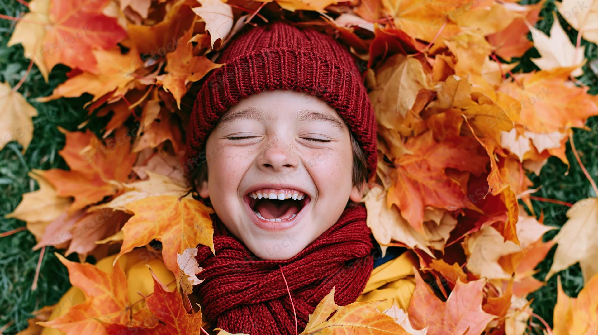 Young boy laughing while lying buried in a pile of autumn maple leaves outdoors