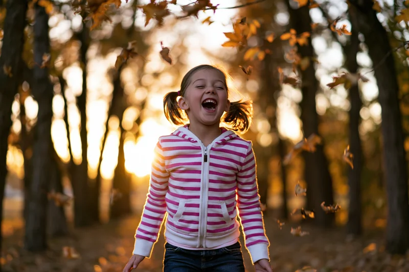 Young girl laughing with mouth open in a forest as autumn leaves fall around her at sunset