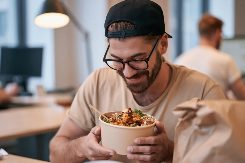 Man eating takeout food from a paper bowl at his office desk