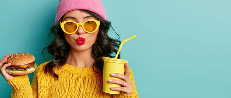 Woman holding burger and yellow soda cup against blue background