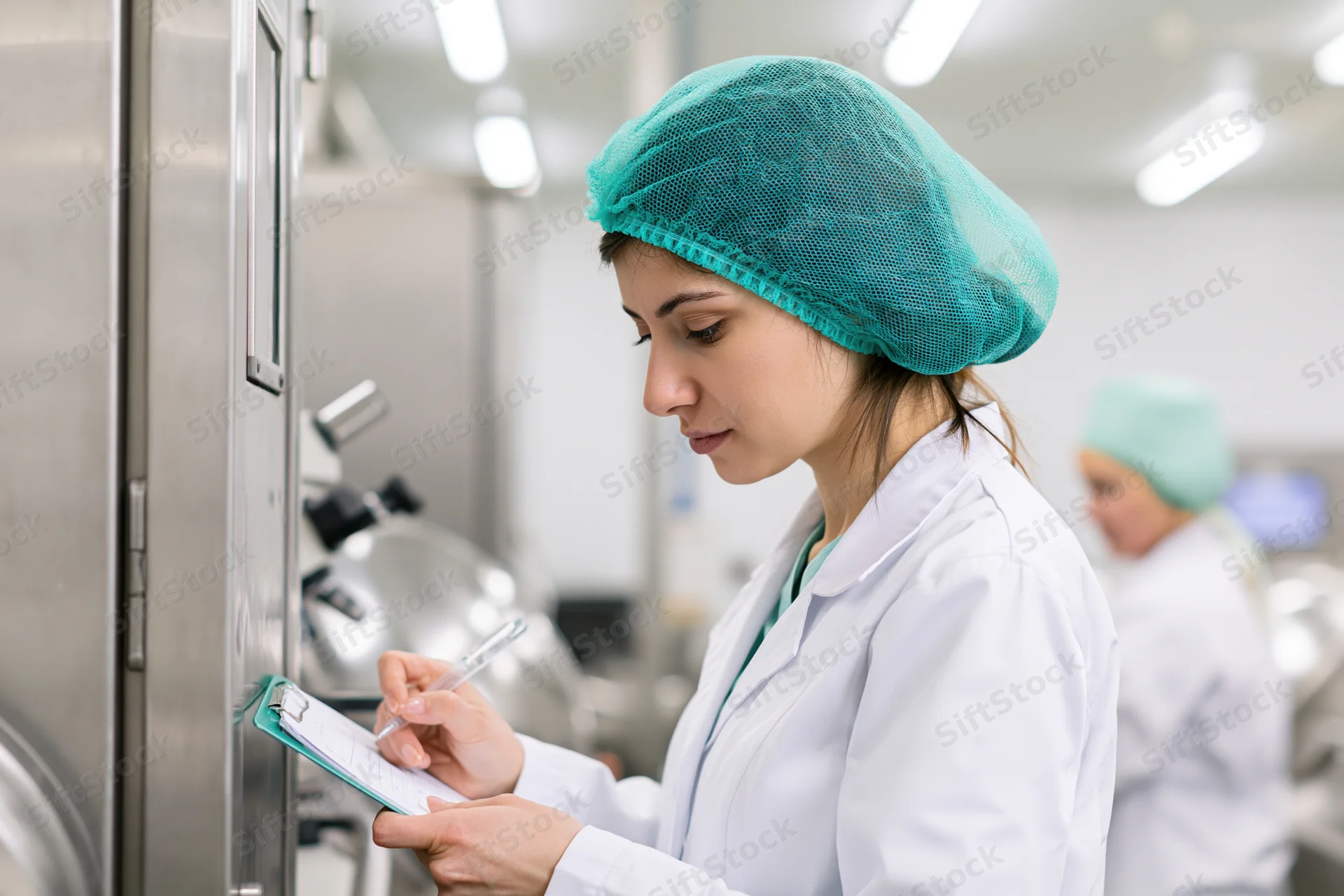 Woman in lab coat writing on clipboard in cleanroom facility