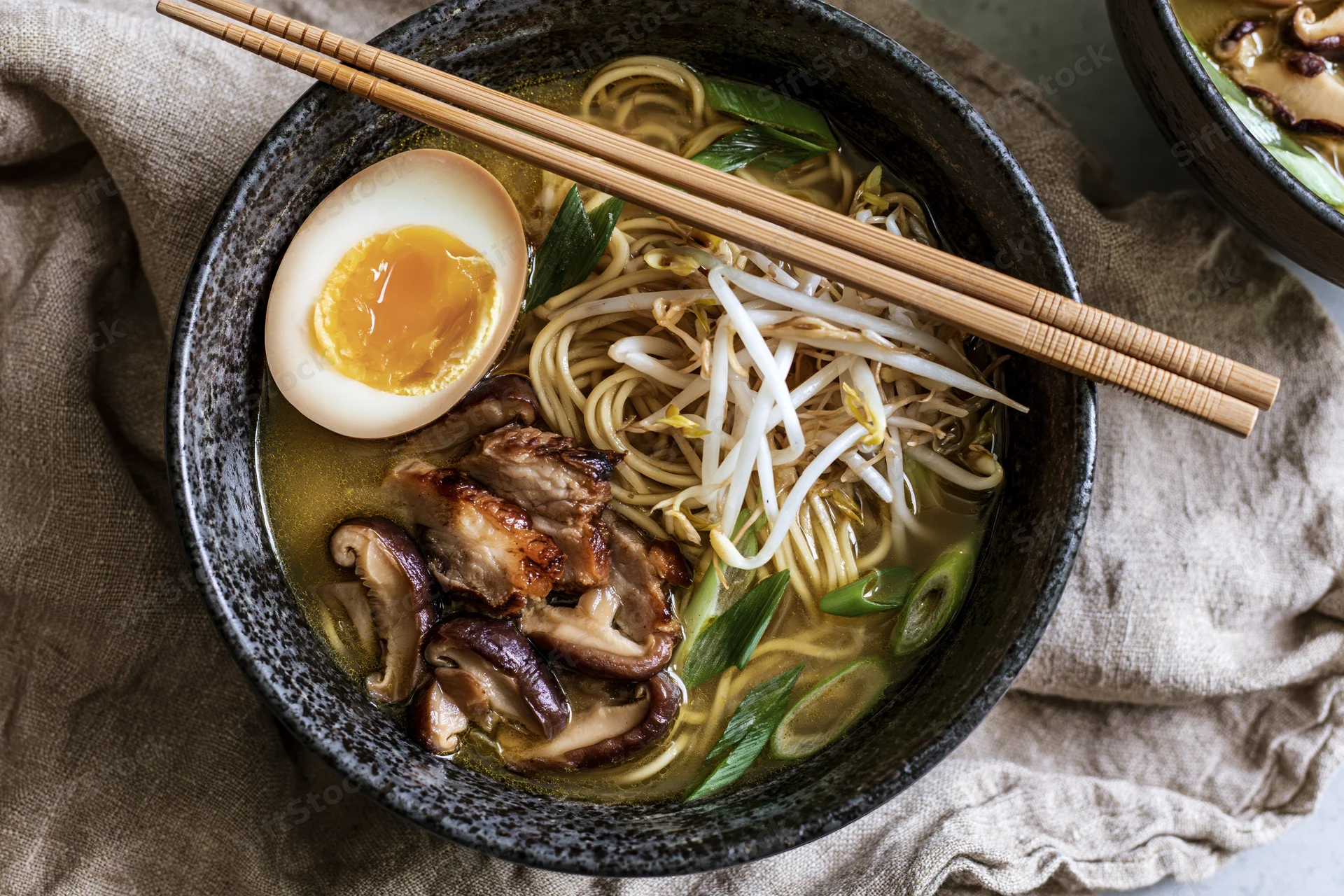 Bowl of Japanese ramen with chopsticks, soft-boiled egg, pork, mushrooms, bean sprouts, and green onions in broth on a linen cloth