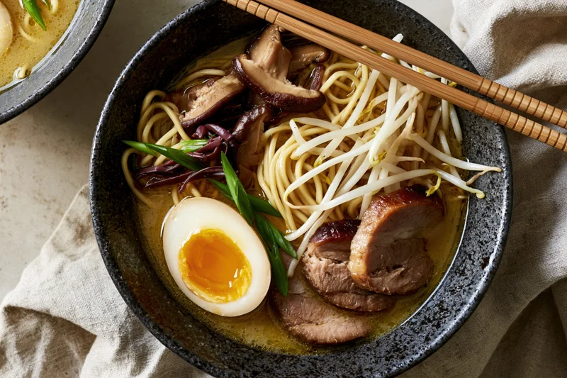 Bowl of Japanese ramen with chopsticks, topped with soft-boiled egg, pork, mushrooms, bean sprouts and green onions, viewed from directly above on a linen cloth