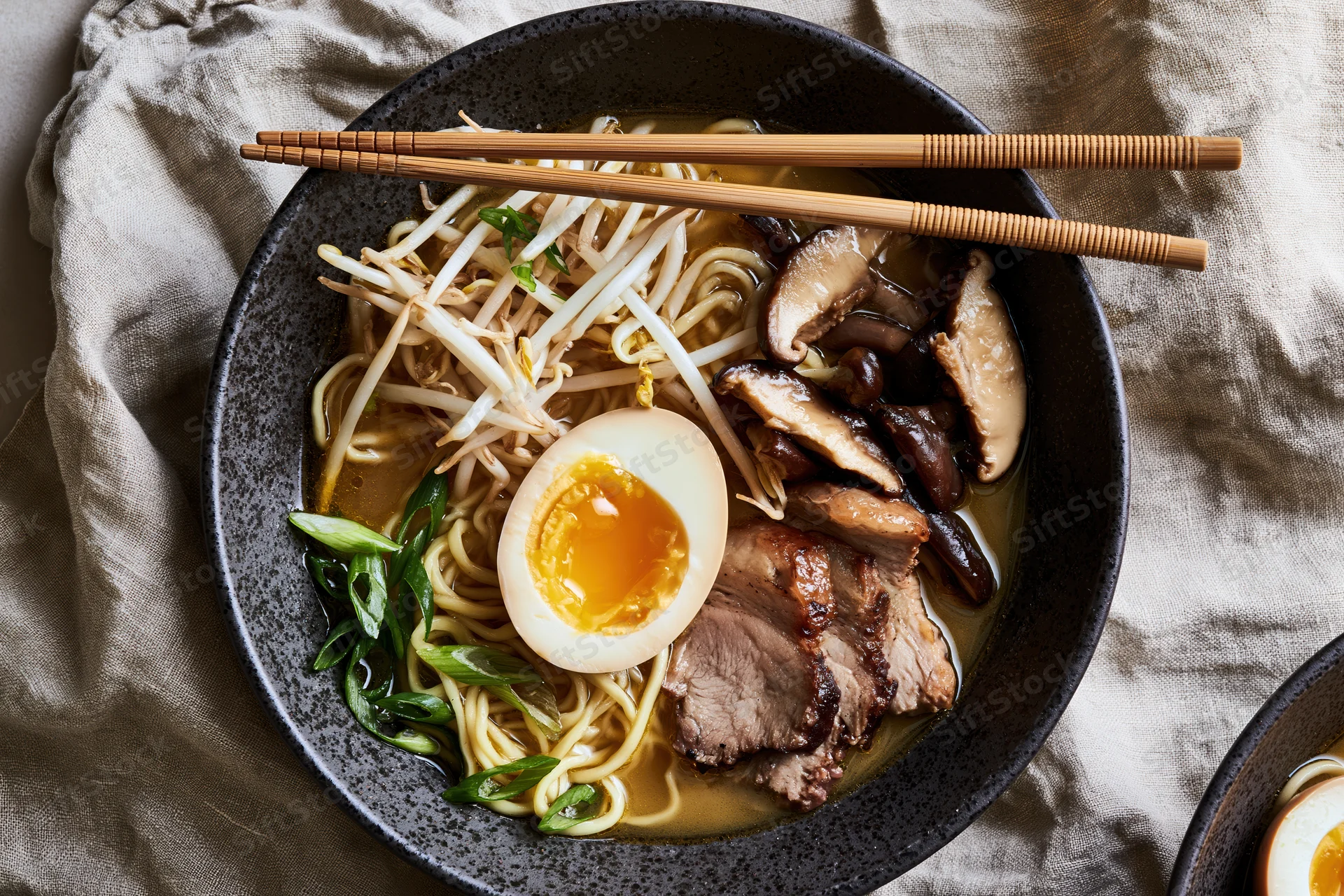 Bowl of japanese ramen with soft-boiled egg, mushrooms, pork, bean sprouts and green onions, overhead view on linen cloth