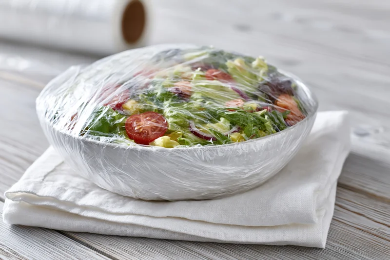 Bowl of fresh vegetable salad covered with plastic wrap on a white napkin on wooden table