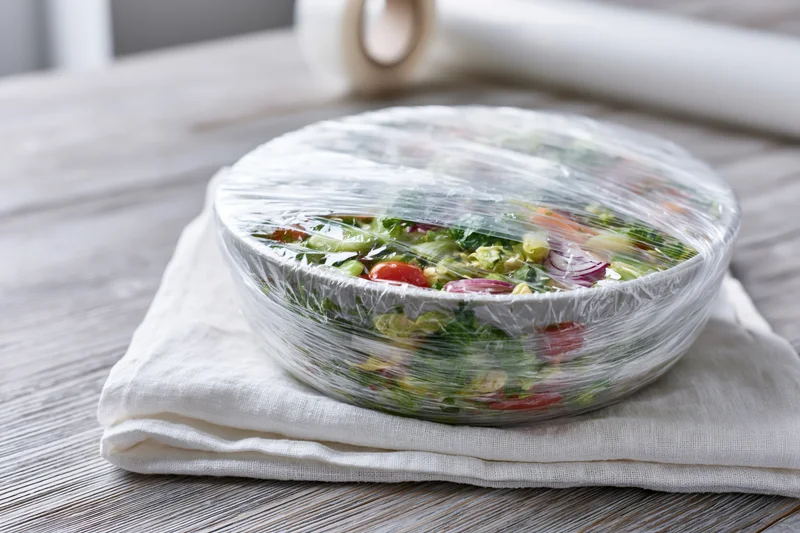 Bowl of fresh vegetable salad covered with plastic wrap on a cloth napkin on wooden table