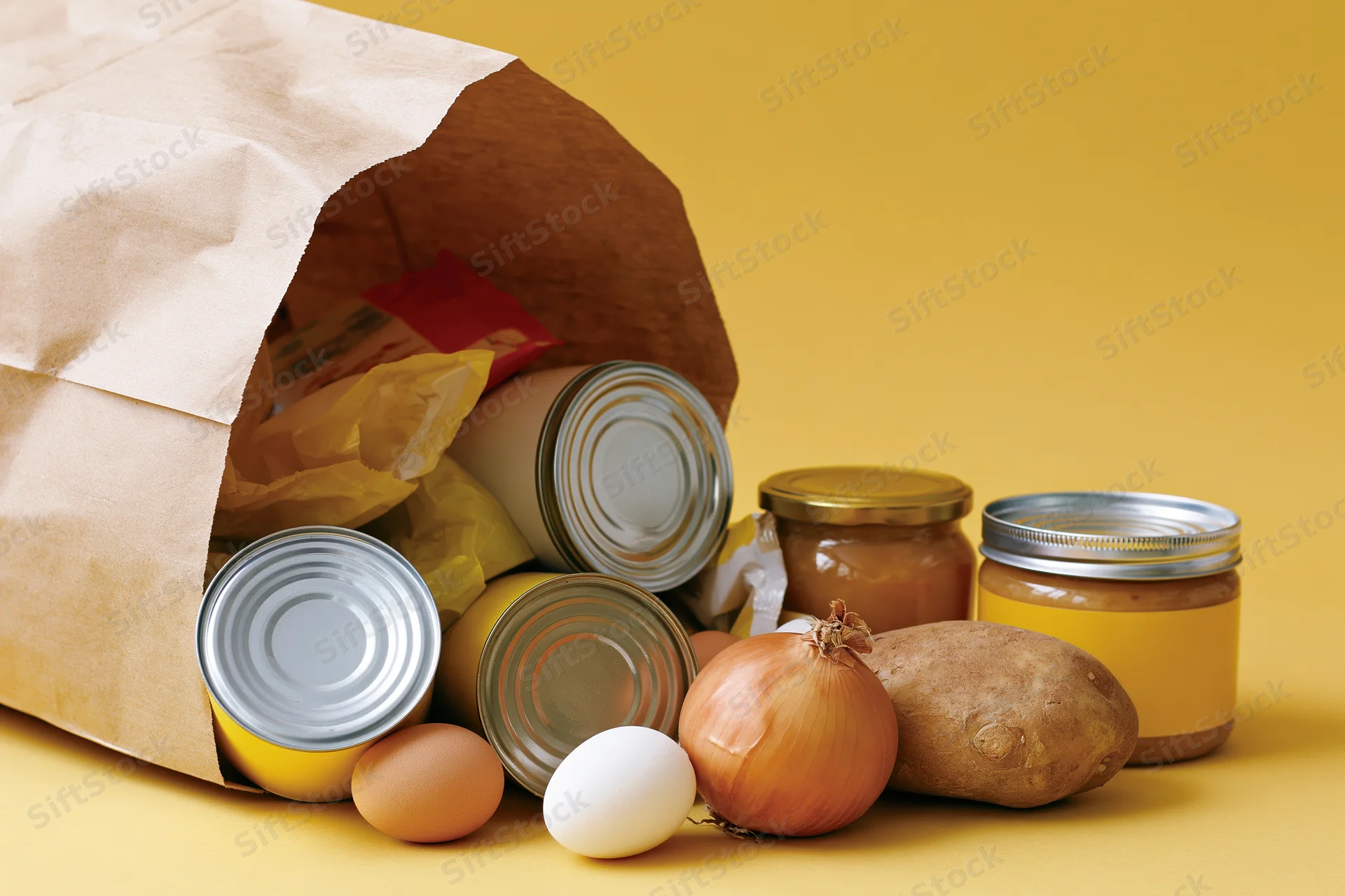 Brown paper grocery bag tipped over spilling canned goods and fresh produce on yellow surface