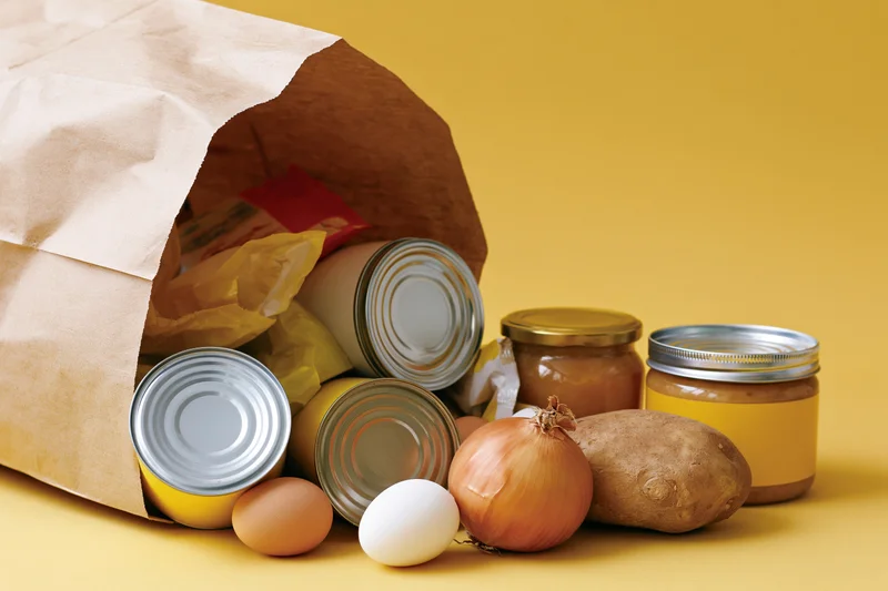 Brown paper grocery bag tipped over spilling canned goods and fresh produce on yellow surface