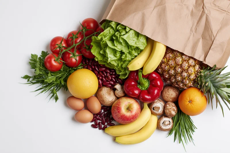 Brown paper grocery bag tipped over spilling fresh fruits and vegetables onto white surface