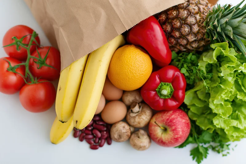 Brown paper grocery bag tipped over spilling fresh fruits and vegetables onto white surface