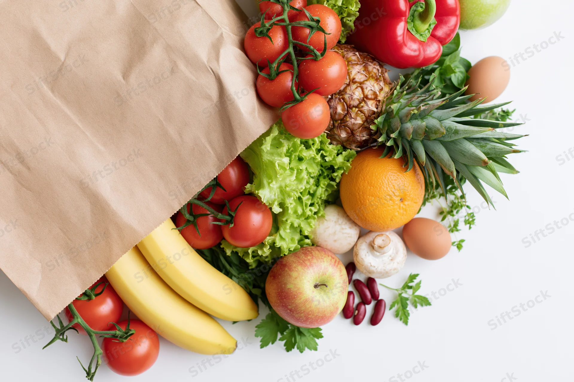 Brown paper grocery bag tipped over with fresh fruits and vegetables spilling out onto white surface