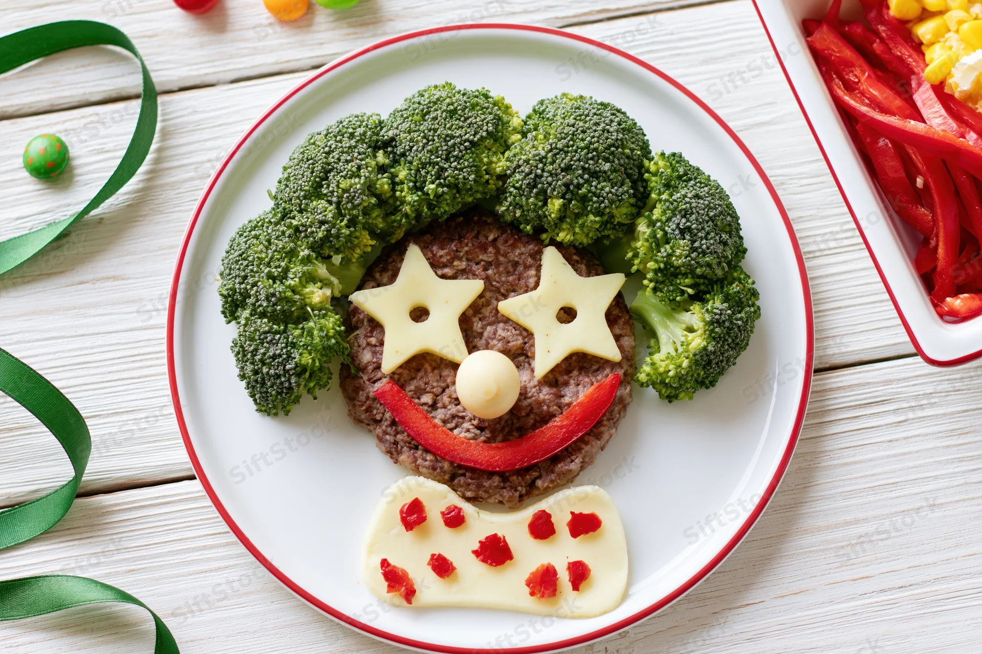Clown face made from broccoli, cheese, meat and pepper on a white plate with red rim, arranged as a childrens meal on a wooden table