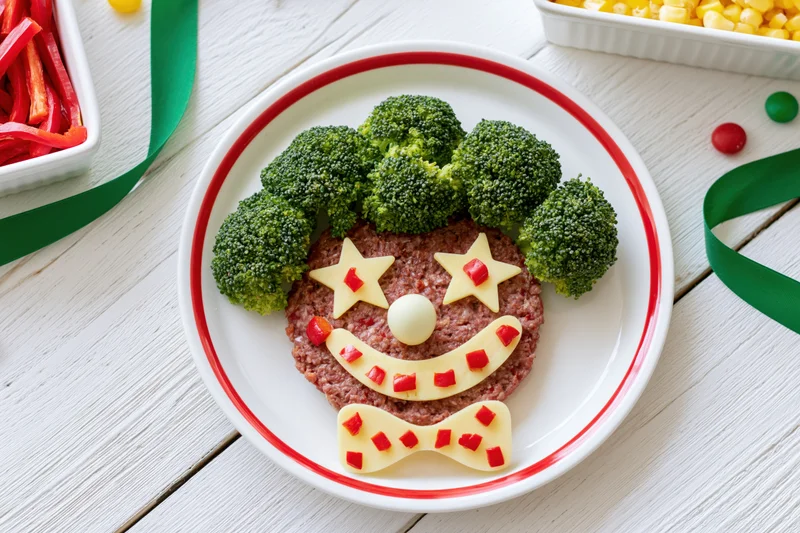 Clown face made from broccoli, hamburger, cheese and red pepper on a white round plate on a wooden table