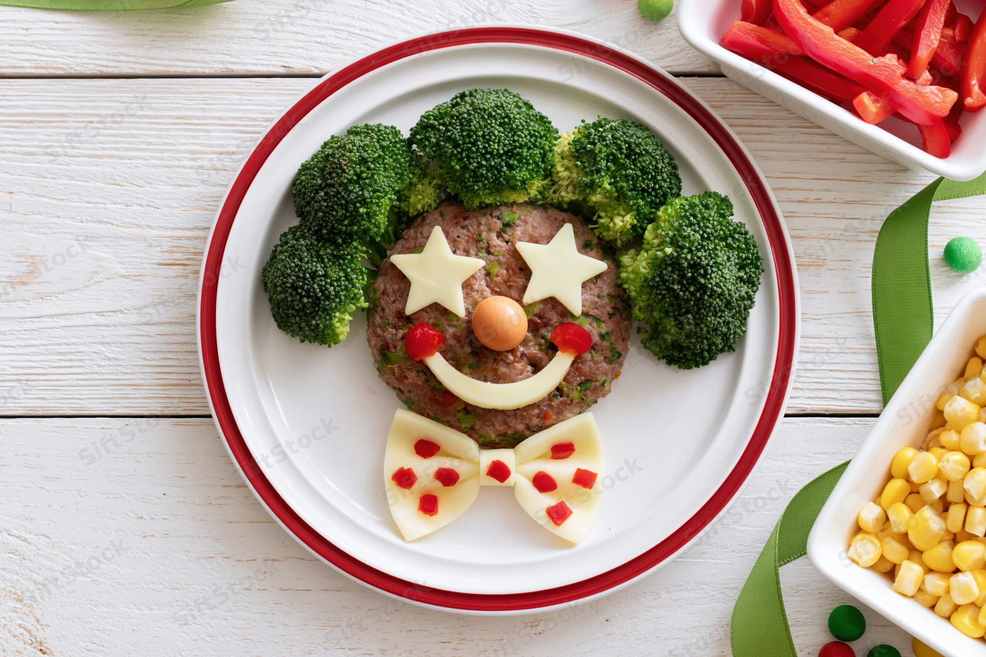 Clown face made from broccoli, cheese, meat and vegetables on a white plate with red rim, surrounded by side dishes on a wooden table