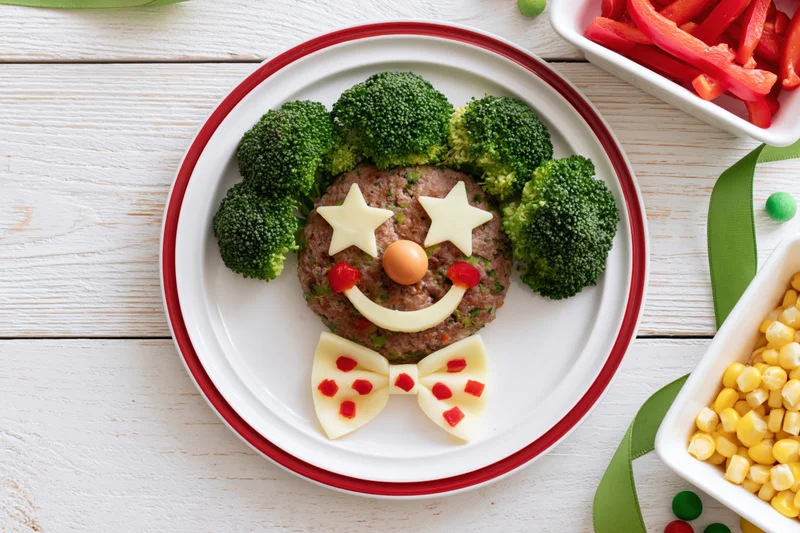 Clown face made from broccoli, cheese, meat and vegetables on a white plate with red rim, surrounded by side dishes on a wooden table