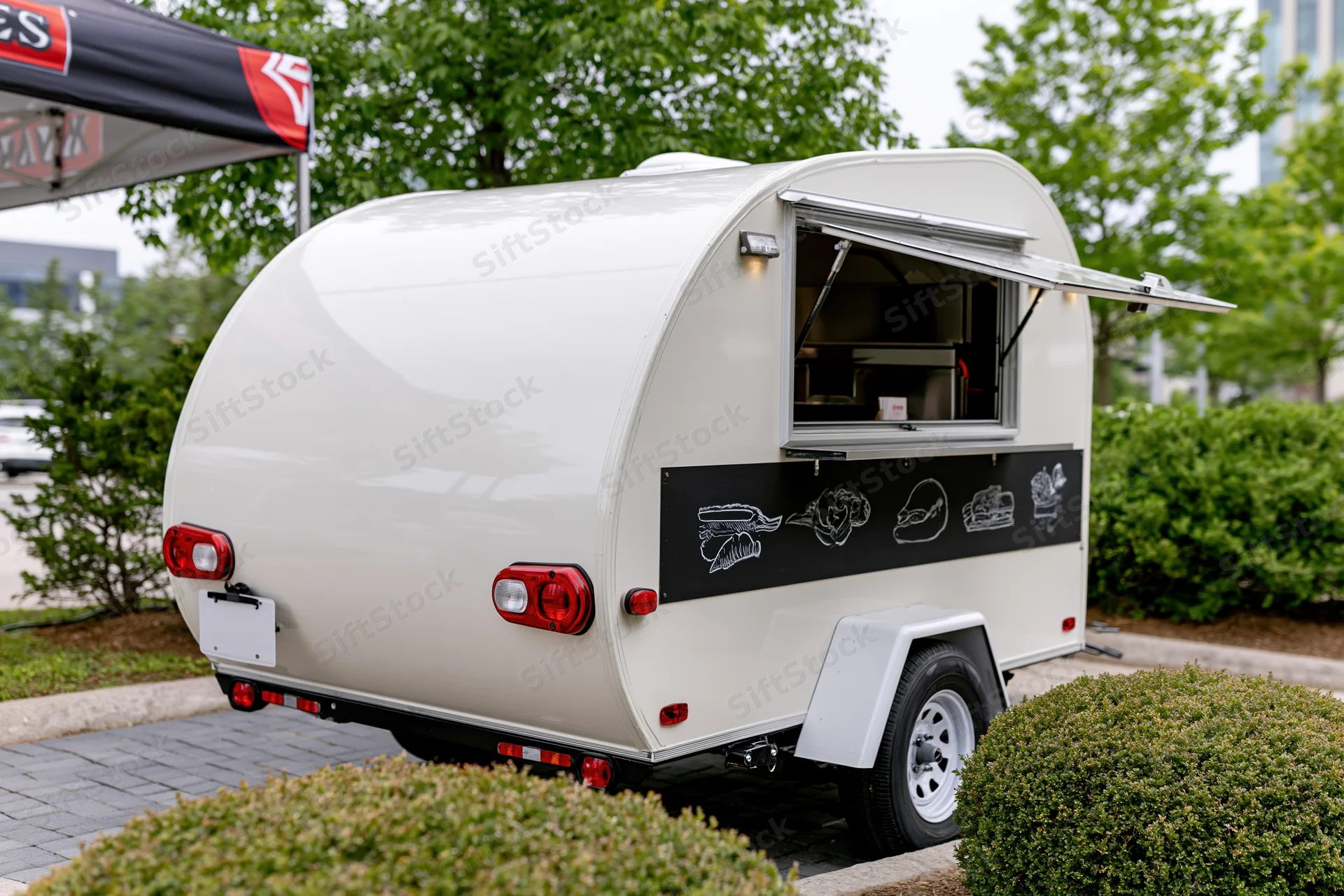 White compact food trailer with open hinged service window parked outdoors near greenery