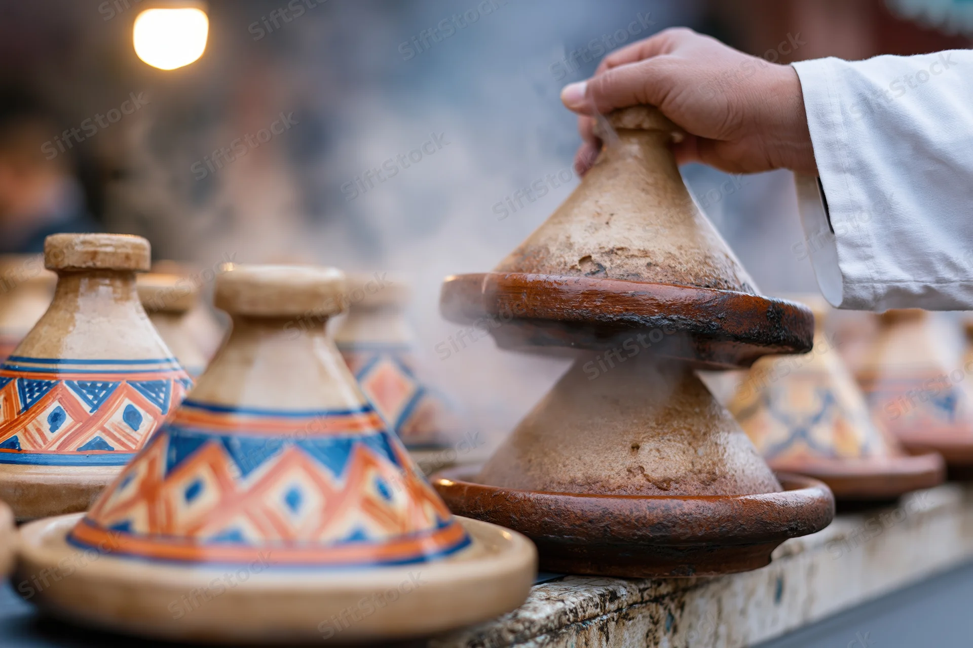Hand lifting conical lid from steaming moroccan tagine at outdoor market stall