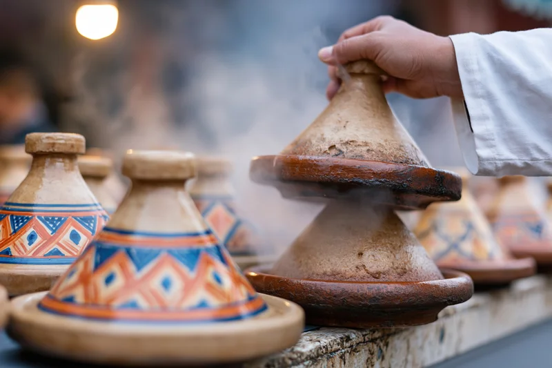Hand lifting conical lid from steaming moroccan tagine at outdoor market stall