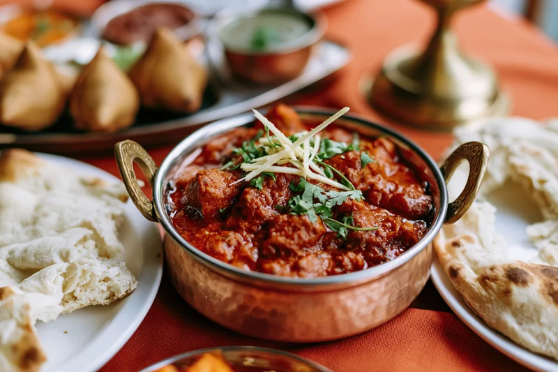 Copper karahi bowl filled with red curry garnished with cilantro and onion strips on a table with naan and samosas