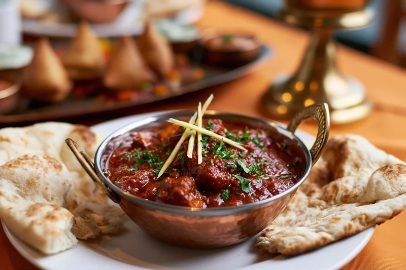 Copper karahi bowl filled with red curry garnished with ginger and cilantro, served with naan bread on a white plate at a dining table