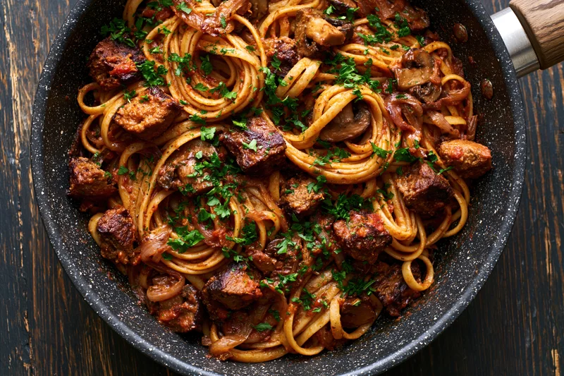 Linguine with meat and mushrooms in a dark speckled frying pan on a wooden surface