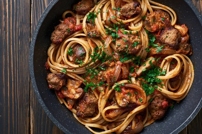 Linguine with meatballs and tomato sauce in a dark speckled frying pan on wooden surface