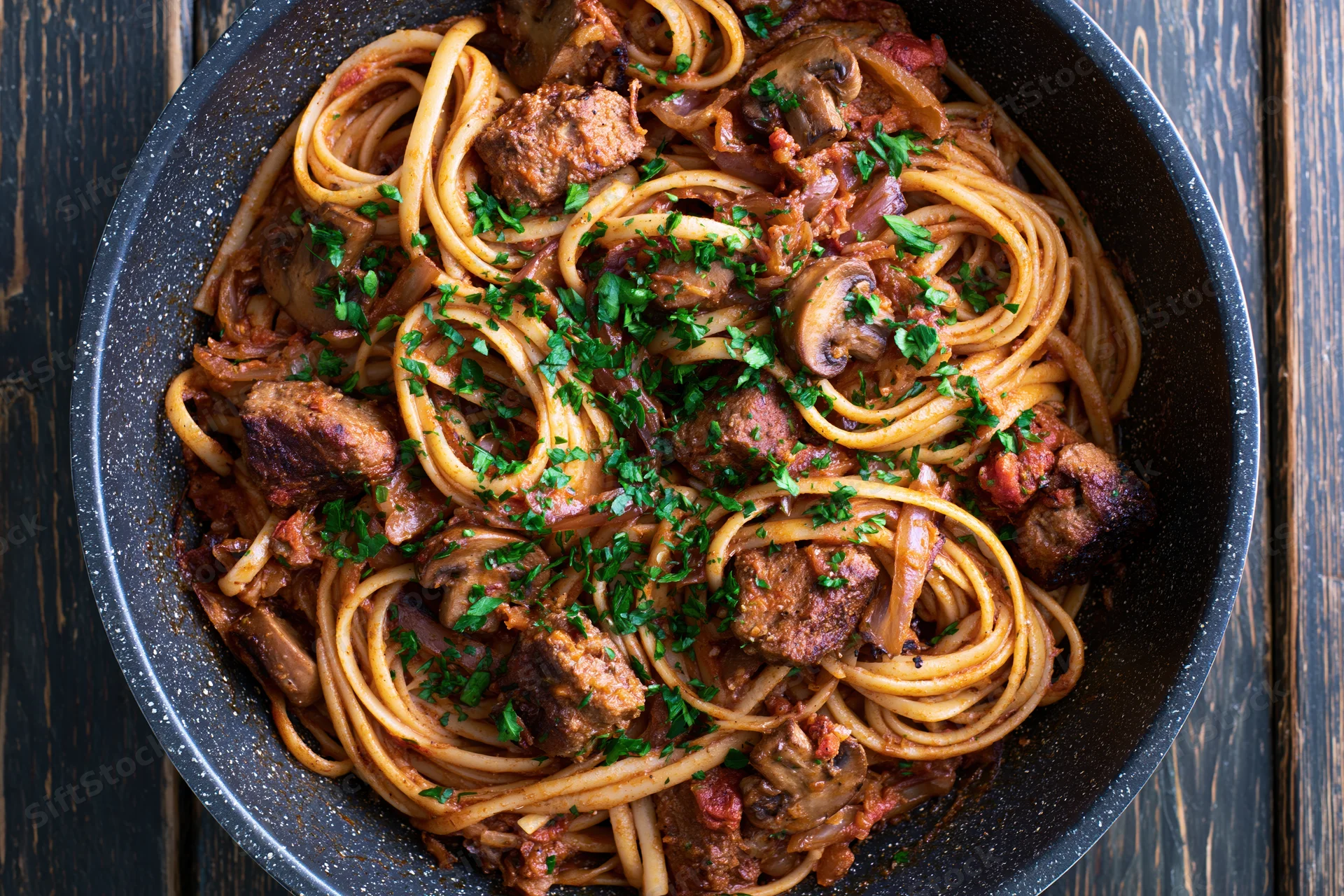Linguine with meat and mushrooms in a dark speckled frying pan on a wooden table