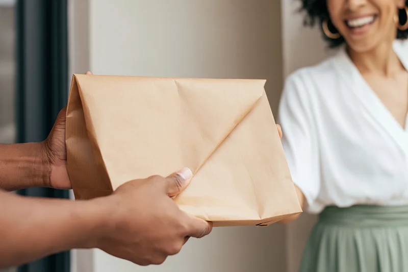 Delivery person handing a folded brown paper bag to a smiling woman at the door