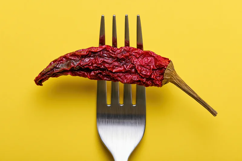 Dried red chili pepper resting on fork tines against yellow background