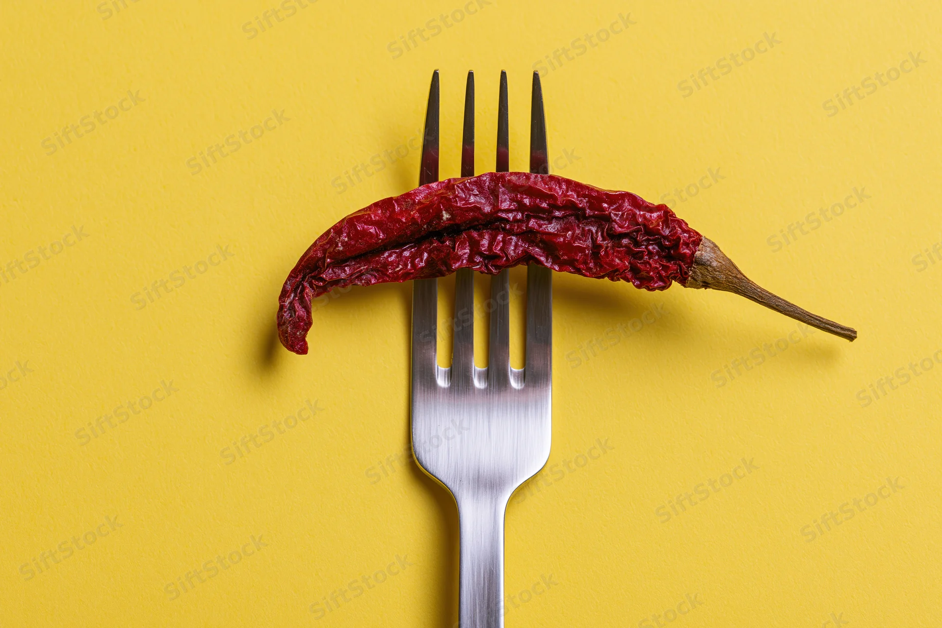 Dried red chili pepper resting across tines of a fork on yellow background