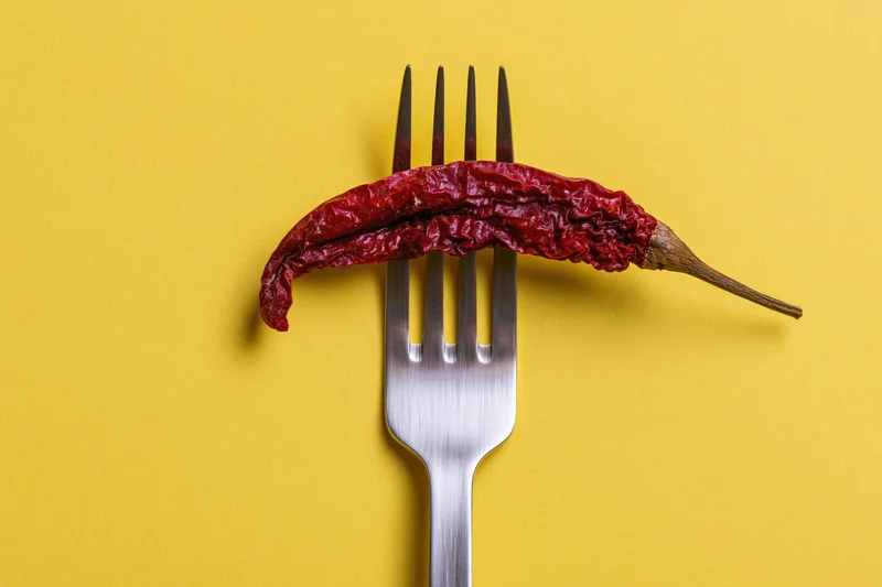 Dried red chili pepper resting across tines of a fork on yellow background