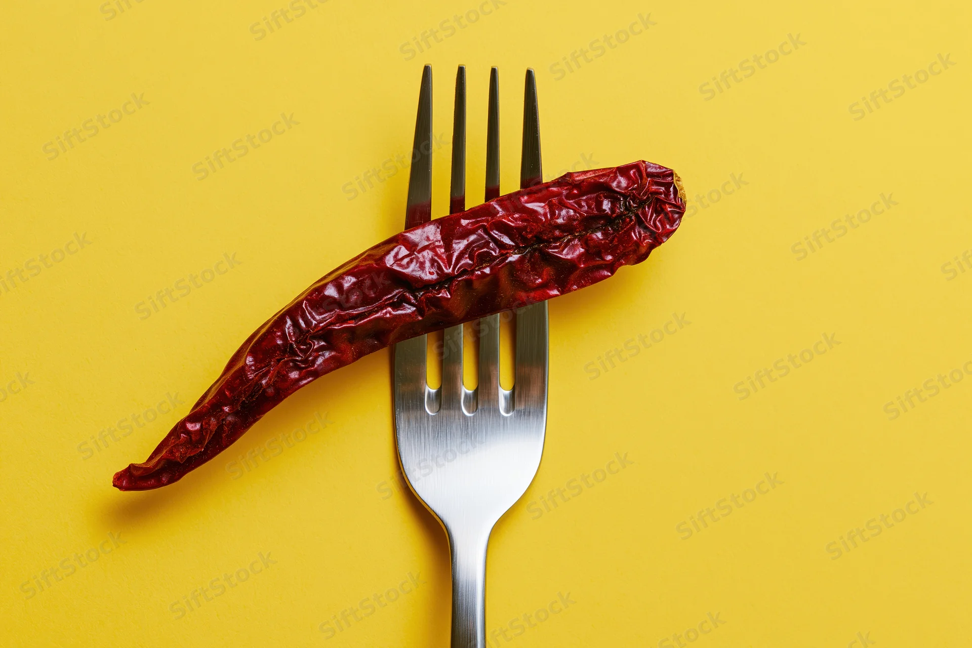 Dried red chili pepper resting across tines of silver fork on yellow background