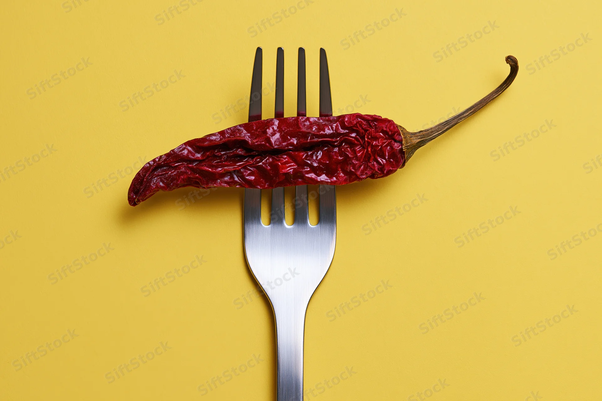 Dried red chili pepper resting across tines of metal fork on yellow background