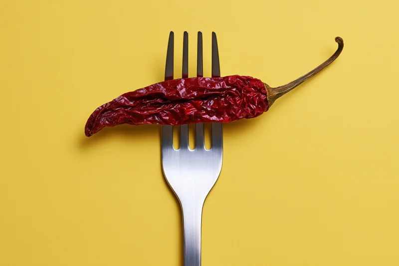 Dried red chili pepper resting across tines of metal fork on yellow background