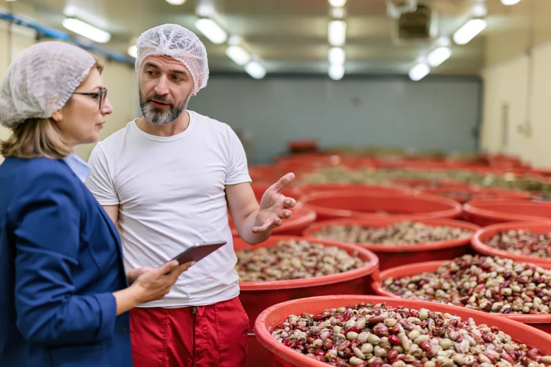 Woman in blue blazer and hairnet discussing coffee beans with man in factory, holding tablet during quality inspection