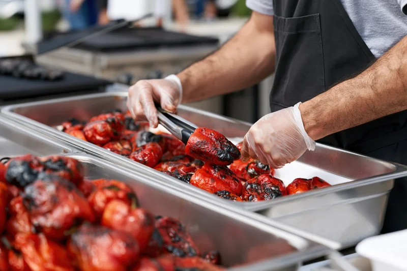 Food service worker using tongs to serve grilled red peppers from metal trays at an outdoor catering event