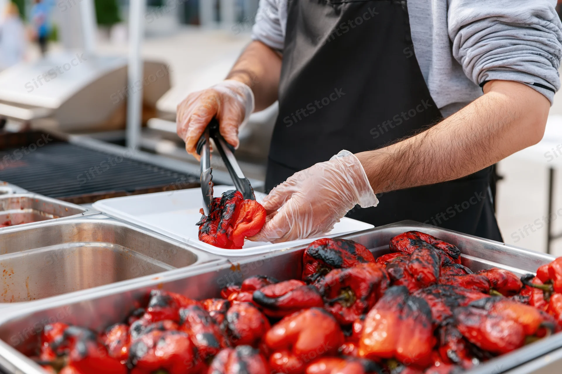 Man in black apron and gloves using tongs to serve grilled red peppers from metal tray at outdoor food stall