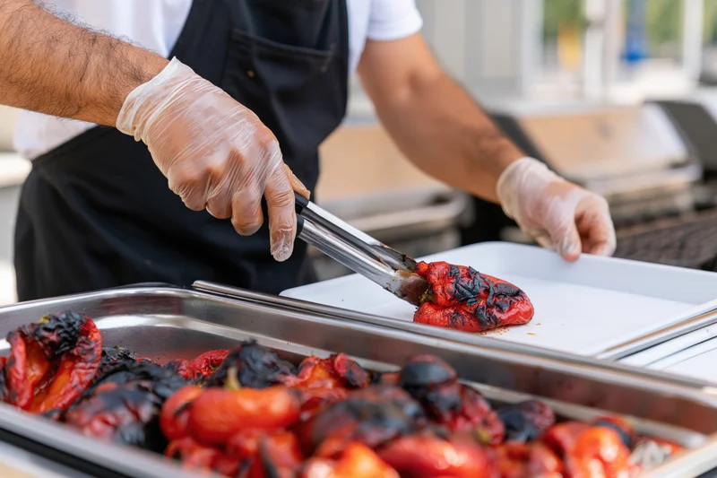 Chef using tongs to transfer roasted red peppers from metal tray to white plate in outdoor kitchen setting