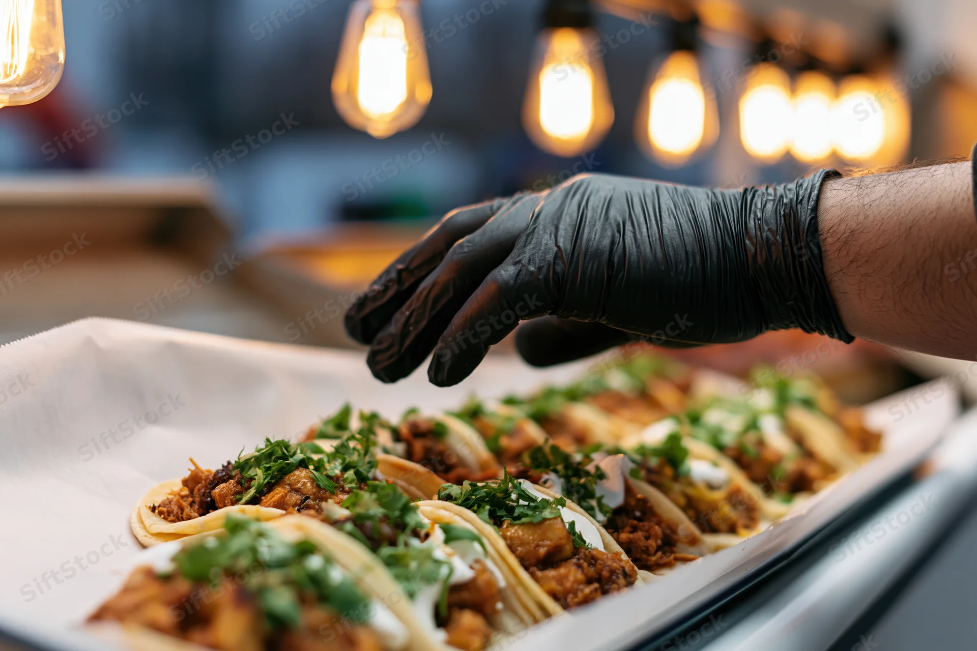Vendor's hand in black nitrile glove reaching to serve tacos on parchment-lined tray in restaurant kitchen