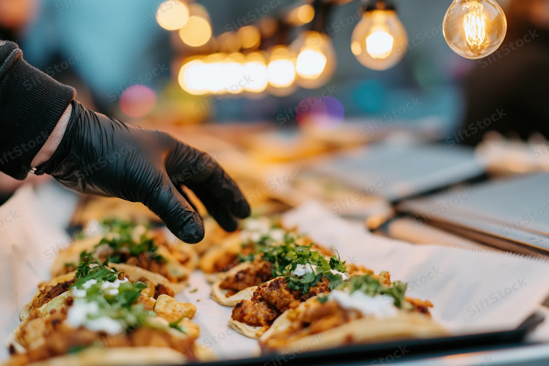 Vendor's hand in black nitrile glove reaching to serve tacos on a tray at a street food market