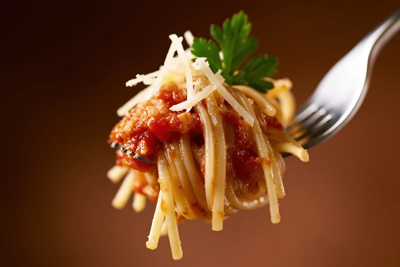Fork twirling spaghetti with tomato sauce and grated cheese, garnished with parsley, against a warm brown background
